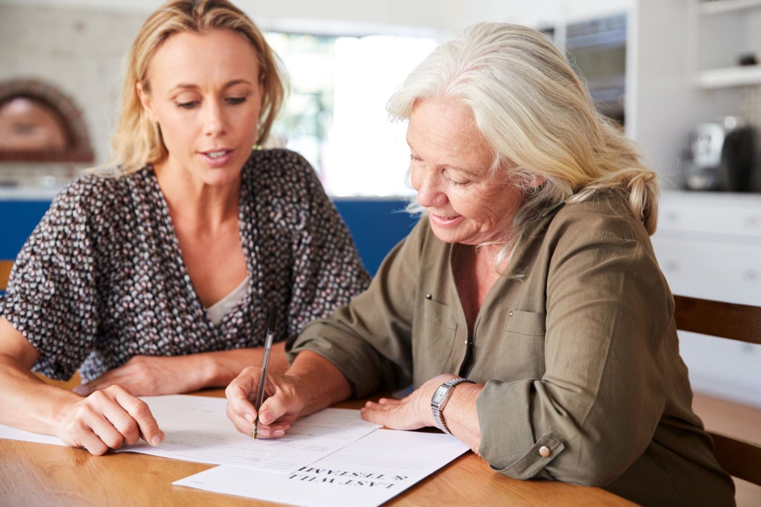 Bildet viser en eldre kvinne som signerer et papir, med hjelp fra en yngre kvinne.