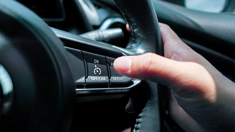Mann som holder en hånd på bilrattet og en finger på cruisekontroll-knappen.