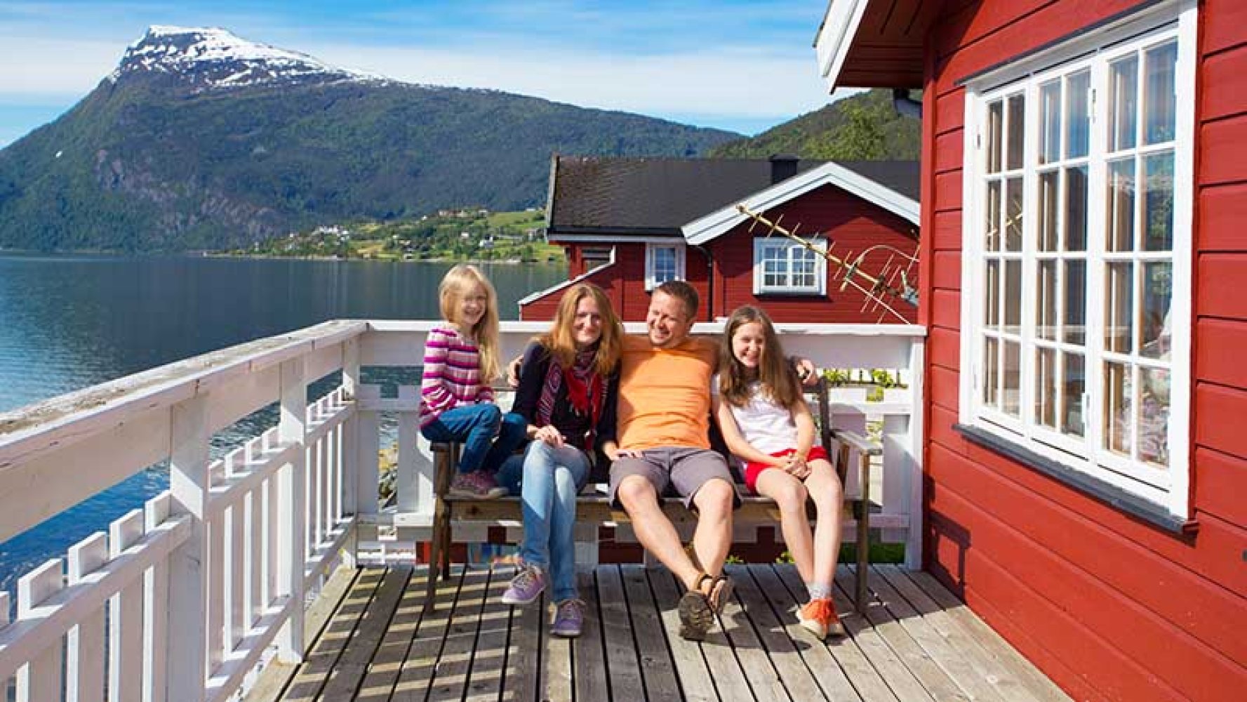 Fornøyd familie på hytteterrasse med fjord og fjell i bakgrunnen.